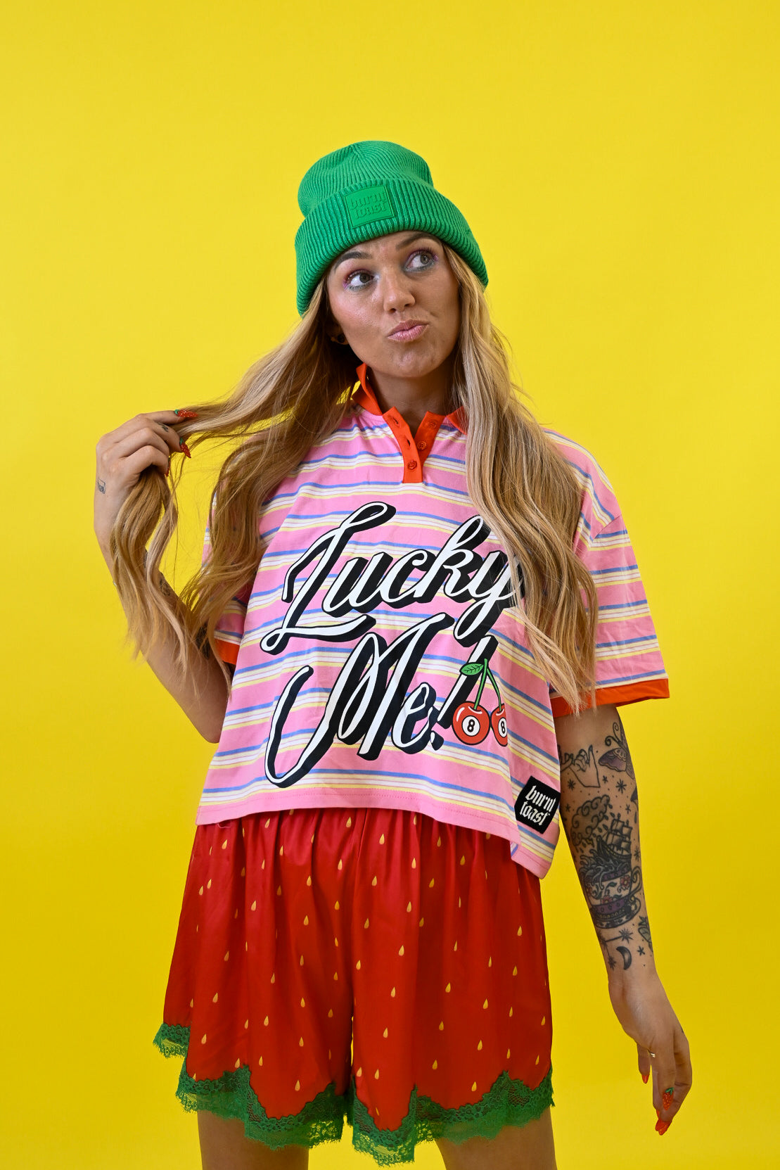 A woman posing in a vibrant studio setting against a solid yellow background, wearing a green beanie, a pink and white striped t-shirt with 'Lucky Me!' text, and red strawberry-patterned shorts