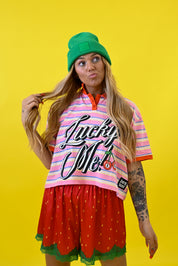 A woman posing in a vibrant studio setting against a solid yellow background, wearing a green beanie, a pink and white striped t-shirt with 'Lucky Me!' text, and red strawberry-patterned shorts