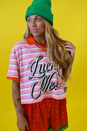 A woman wearing a green beanie, a pink and white striped short-sleeve polo shirt with 'Lucky Me!' text, and a red strawberry-patterned shorts, posed against a solid yellow background