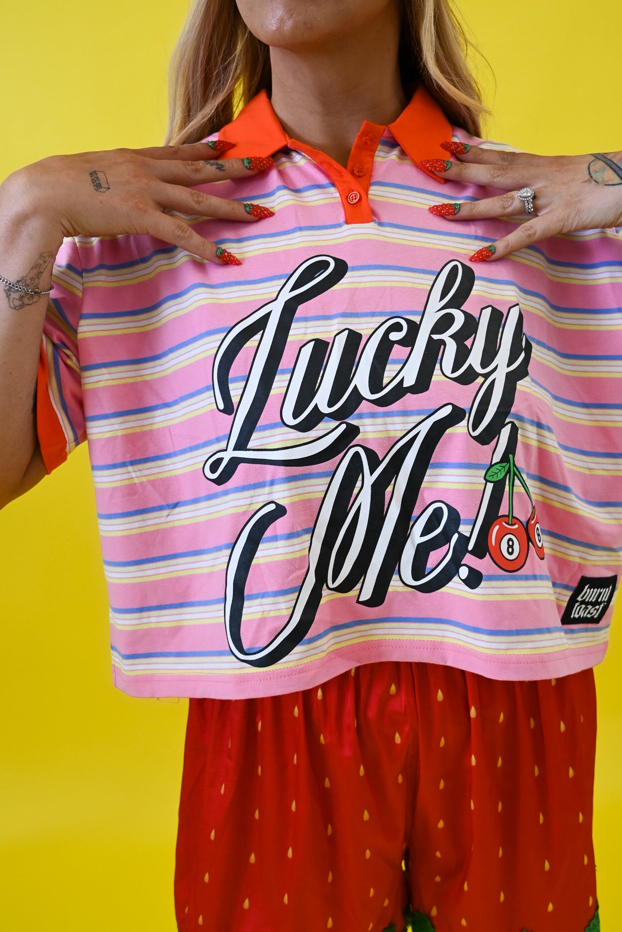 A cropped pink and orange striped polo shirt with 'Lucky Me!' printed in large black script across the front, worn by a model against a bright yellow background