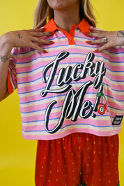 A cropped pink and orange striped polo shirt with 'Lucky Me!' printed in large black script across the front, worn by a model against a bright yellow background
