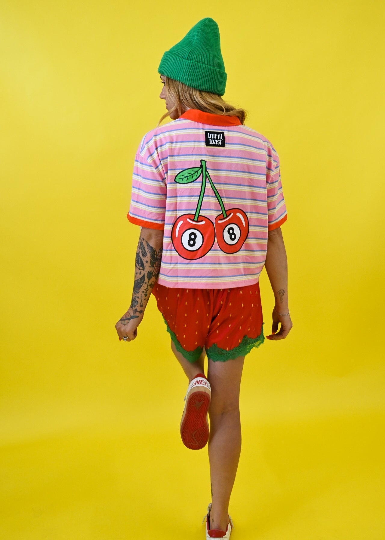 A person shown from behind wearing a pink and white striped t-shirt with a large cherry graphic on the back, red strawberry shorts, a green beanie, and white sneakers, set against a solid yellow background