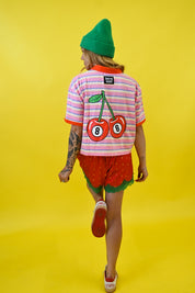 A person shown from behind wearing a pink and white striped t-shirt with a large cherry graphic on the back, red strawberry shorts, a green beanie, and white sneakers, set against a solid yellow background