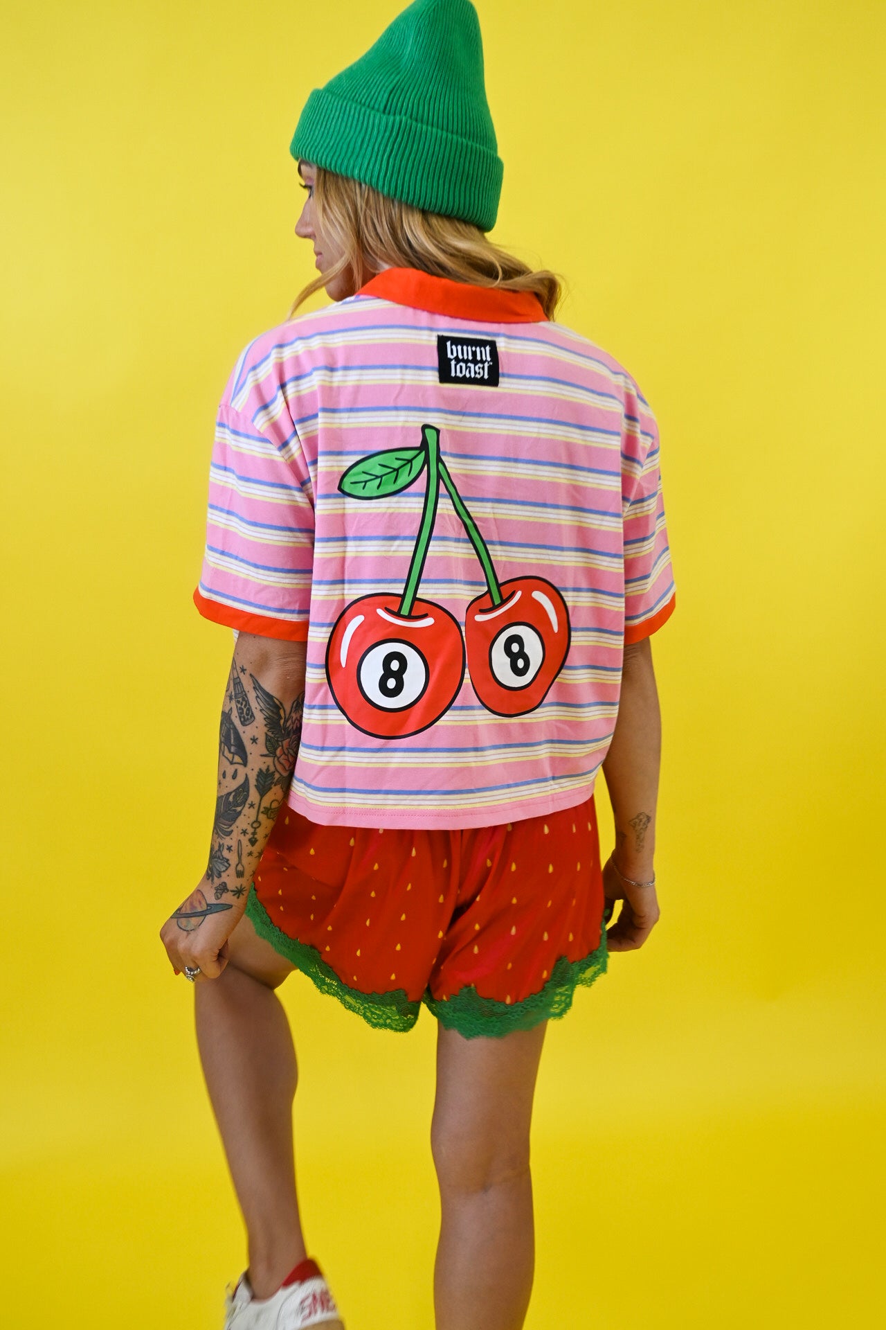 A model from behind wearing a pink and white striped t-shirt with a large cherry graphic on the back, red strawberry shorts, and a green beanie, set against a solid yellow background