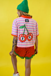 A model from behind wearing a pink and white striped t-shirt with a large cherry graphic on the back, red strawberry shorts, and a green beanie, set against a solid yellow background