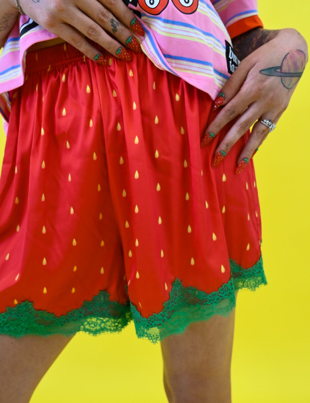 A person wearing bright red strawberry-themed shorts with yellow seed prints and green lace trim, paired with a pink striped shirt against a solid yellow background