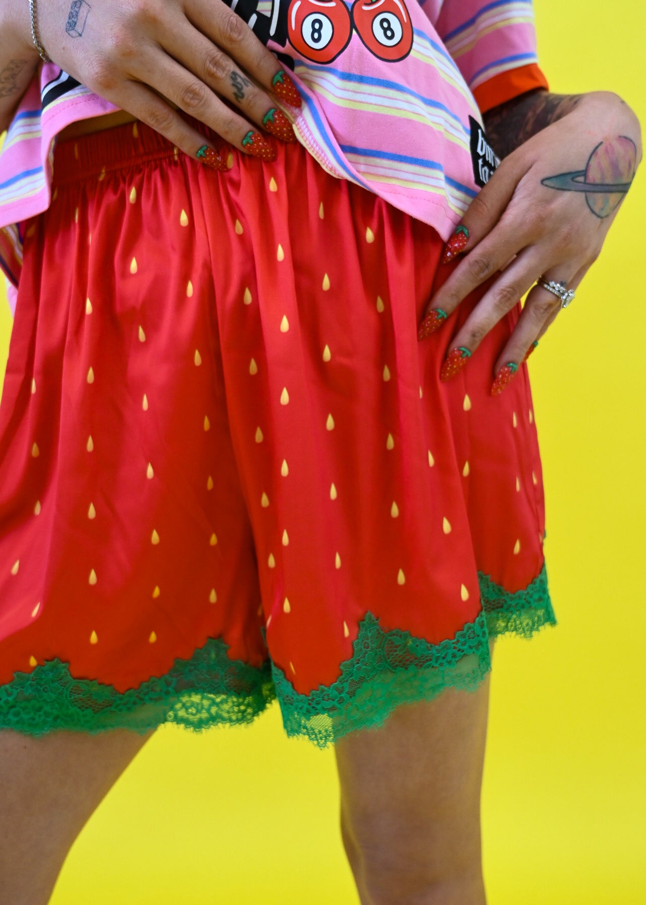 A person wearing bright red strawberry-themed shorts with yellow seed prints and green lace trim, paired with a pink striped shirt against a solid yellow background