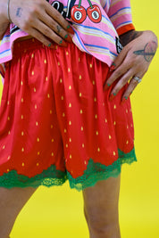 A person wearing bright red strawberry-themed shorts with yellow seed prints and green lace trim, paired with a pink striped shirt against a solid yellow background