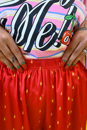 Close-up of a person wearing a pink striped graphic T-shirt with large black script text and a cherry illustration, paired with a bright red skirt patterned with small yellow teardrop shapes. The person's hands are visible at the waist