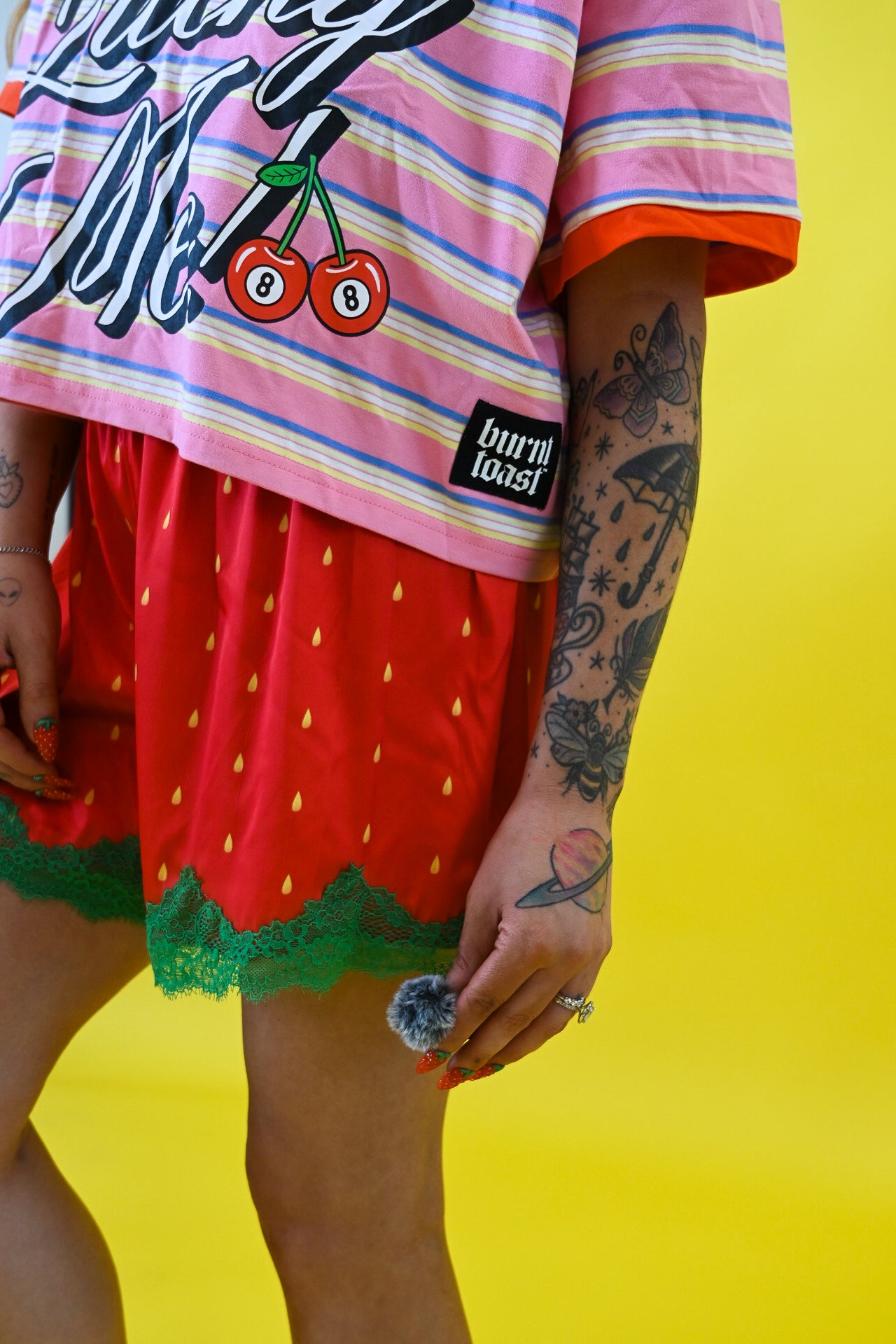 A person wearing a pink and white striped t-shirt with cherry graphics and red strawberry-patterned shorts with green lace trim, posed against a bright yellow background