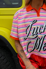 A close-up of a person wearing a pink striped polo shirt with orange collar and cuffs, large black script text reading 'Lucky Me' across the front, and red bottoms, standing beside a bright yellow vehicle