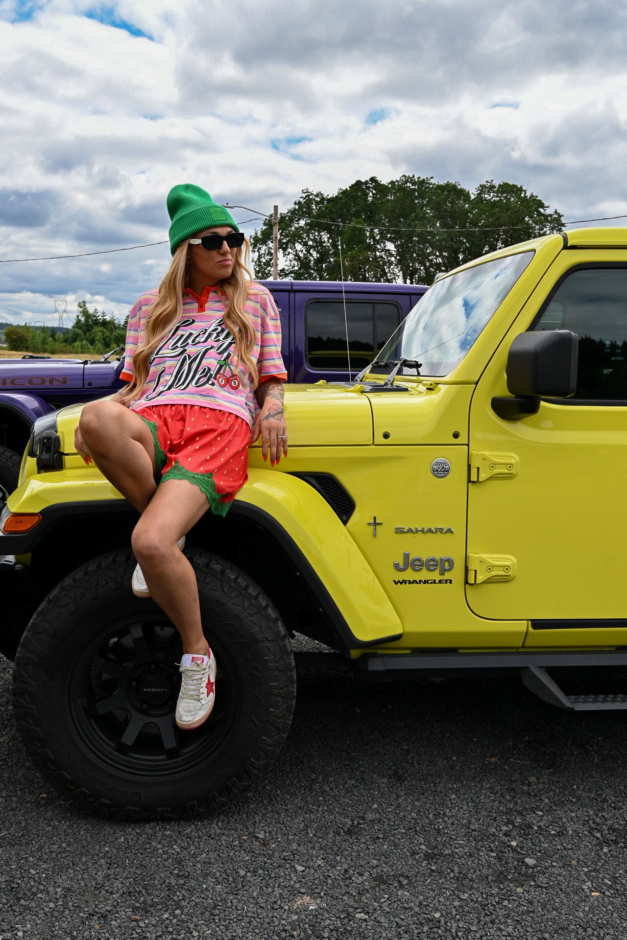 A woman wearing a green beanie, sunglasses, a pink striped shirt, and red shorts sits on the front fender of a bright yellow Jeep. The photo is taken outdoors in a parking lot under a cloudy sky