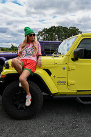 A woman wearing a green beanie, sunglasses, a pink striped shirt, and red shorts sits on the front fender of a bright yellow Jeep. The photo is taken outdoors in a parking lot under a cloudy sky