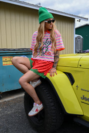 A woman wearing a green beanie, sunglasses, a pink and white striped graphic t-shirt, a red and green skirt, and white sneakers with red stars sits on the tire of a bright yellow vehicle