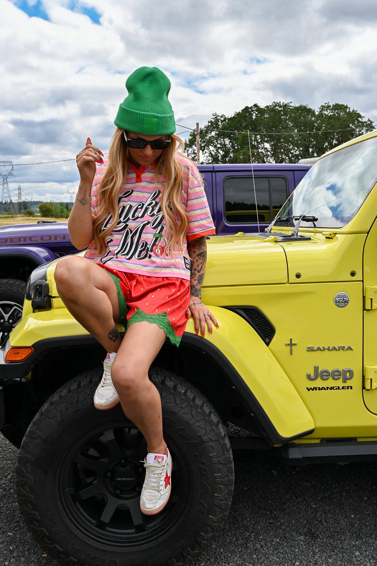 A woman wearing a green beanie, sunglasses, a pink striped t-shirt, and green shorts sits on the front fender of a bright yellow Jeep Wrangler. The photo is taken outdoors in a parking lot with other vehicles and a cloudy sky in the background