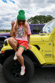 A woman wearing a green beanie, sunglasses, a pink striped t-shirt, and green shorts sits on the front fender of a bright yellow Jeep Wrangler. The photo is taken outdoors in a parking lot with other vehicles and a cloudy sky in the background