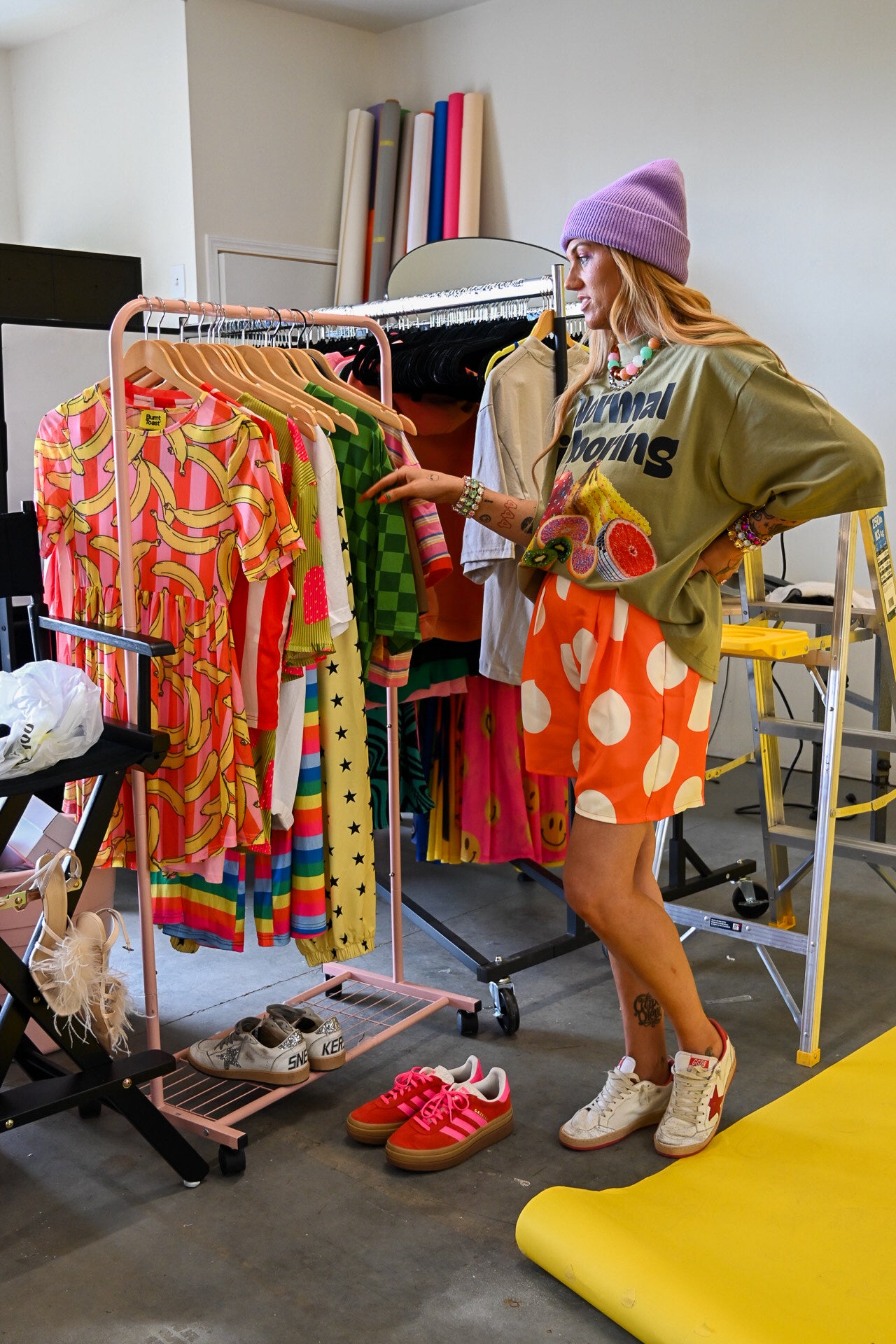 A woman stands in a clothing studio wearing an oversized olive green graphic shirt, a bright orange and white polka dot skirt, white sneakers, and a lavender beanie. She is gesturing toward a pink clothing rack filled with colorful patterned clothing