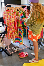 A woman in an olive green shirt and orange polka-dot skirt browses a pink clothing rack filled with colorful patterned dresses in a retail store. She is wearing red and white sneakers