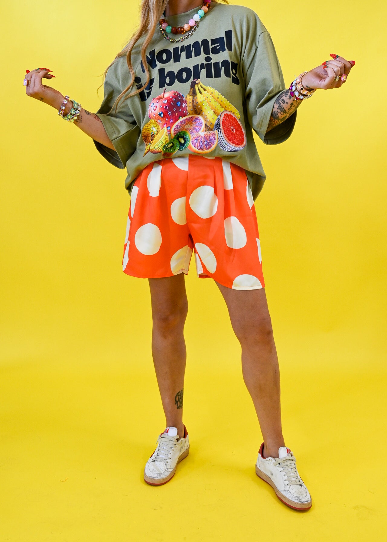 A model wearing a green graphic t-shirt with fruit illustrations, orange shorts with large white polka dots, and white sneakers, posed against a solid bright yellow background