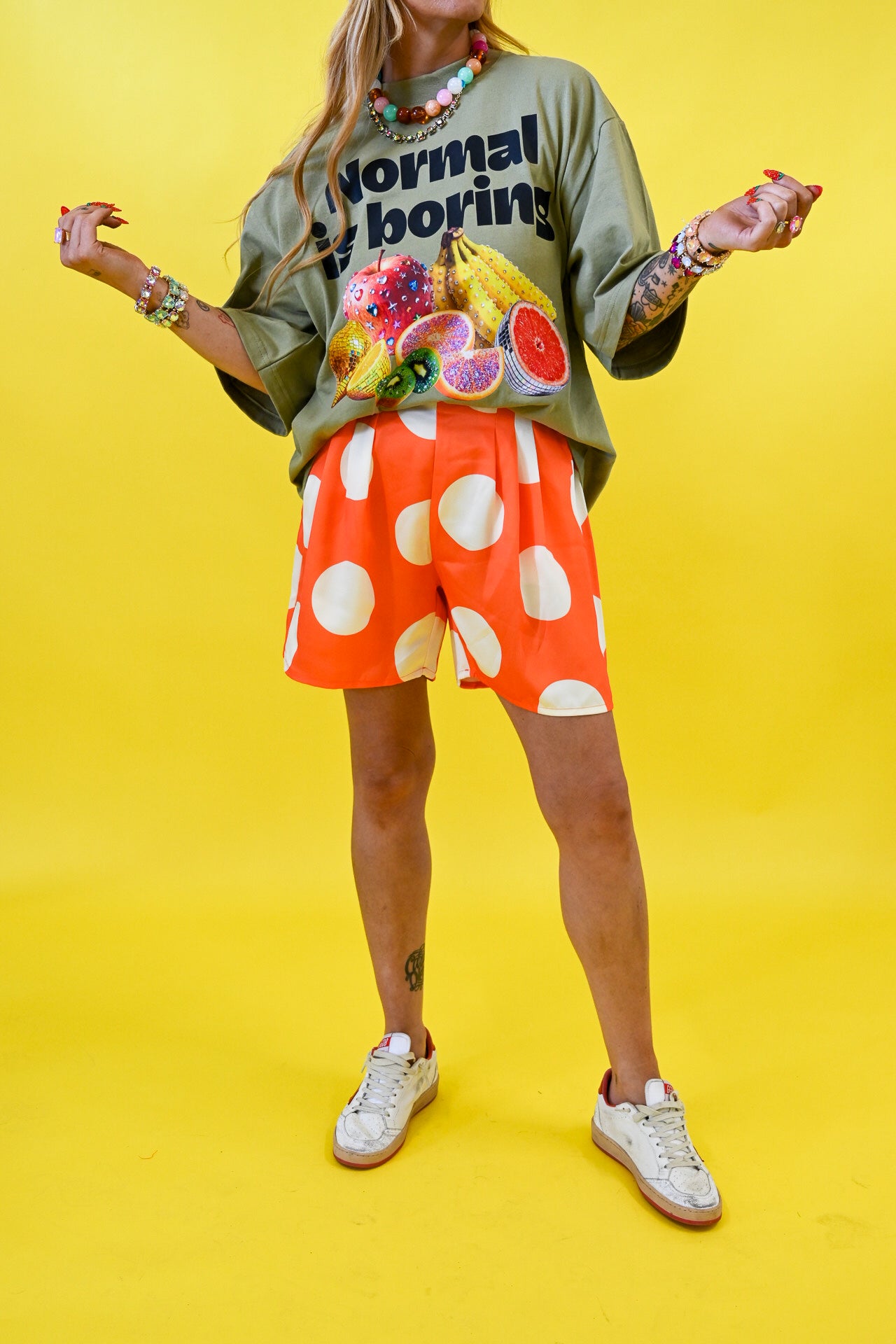 A model wearing a green graphic t-shirt with fruit illustrations, orange shorts with large white polka dots, and white sneakers, posed against a solid bright yellow background