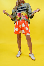 A model wearing a green graphic t-shirt with fruit illustrations, orange shorts with large white polka dots, and white sneakers, posed against a solid bright yellow background