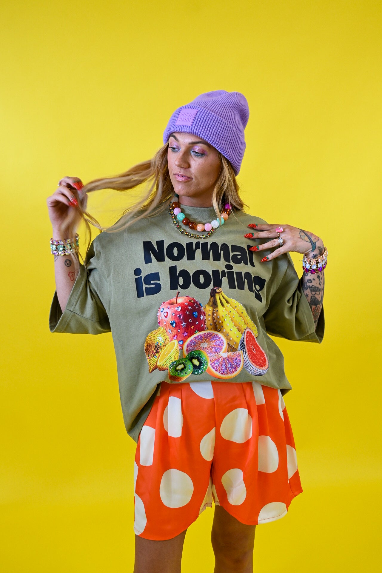 A woman modeling a graphic t-shirt that reads 'Normal is boring' above a fruit illustration, paired with orange shorts featuring white polka dots and a purple beanie, set against a solid yellow background 