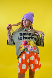 A woman modeling a graphic t-shirt that reads 'Normal is boring' above a fruit illustration, paired with orange shorts featuring white polka dots and a purple beanie, set against a solid yellow background 