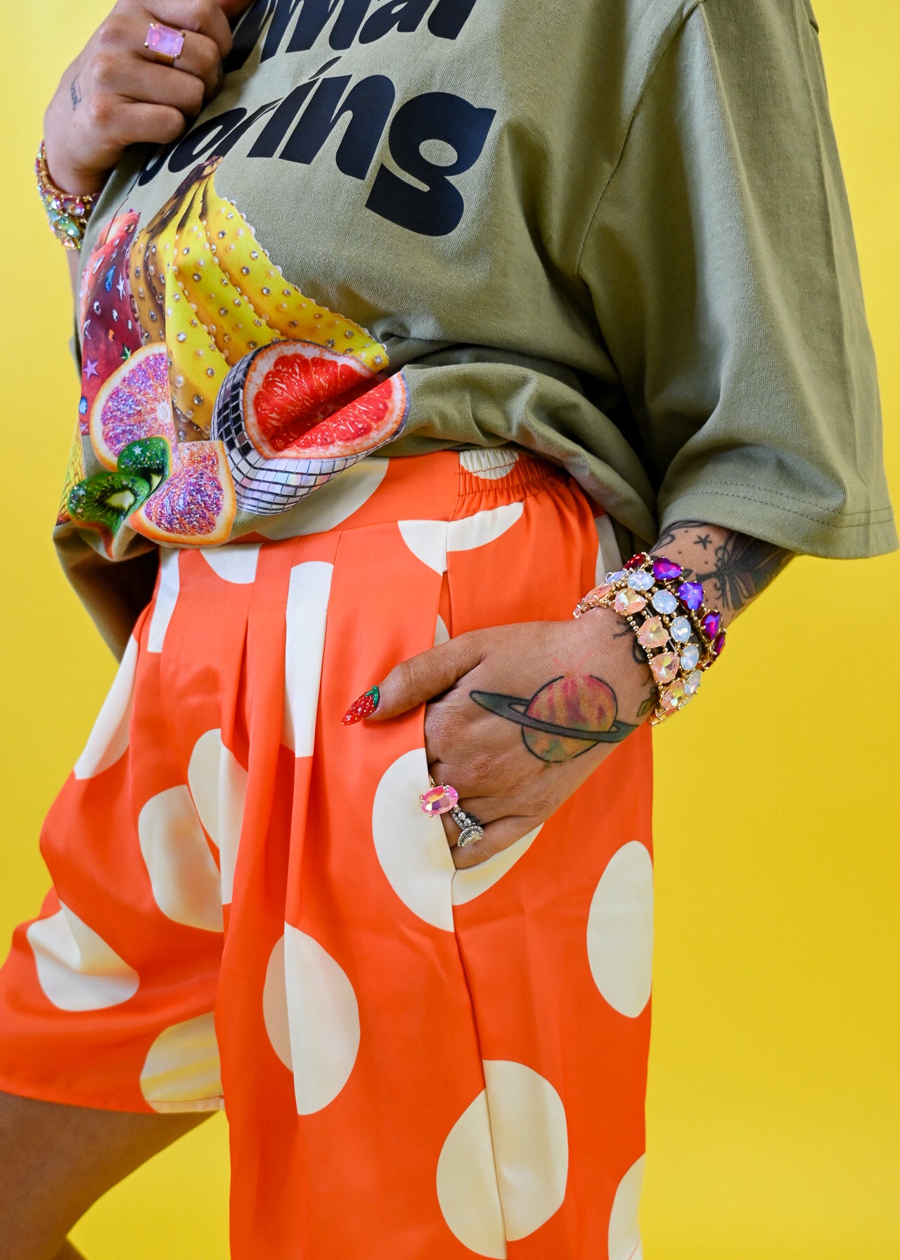 A person wearing a loose olive green graphic T-shirt and bright orange shorts with large white polka dots, posed against a solid yellow background, hand in pocket, close up view