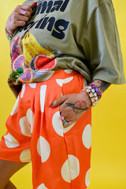 A person wearing a loose olive green graphic T-shirt and bright orange shorts with large white polka dots, posed against a solid yellow background, hand in pocket, close up view