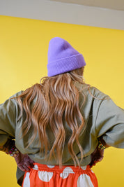 A person shown from behind wearing a lavender knit beanie, an olive green shirt, and orange-and-white patterned shorts against a solid yellow wall 