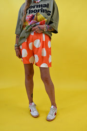 A person wearing bright orange shorts with large white polka dots, an olive green graphic T-shirt, and white sneakers, standing against a solid yellow studio background
