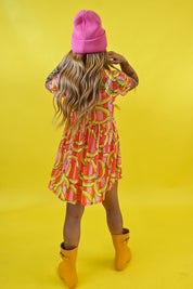 A model shown from behind wearing a pink beanie, a short-sleeve dress with a vibrant orange, pink, and yellow abstract pattern, and yellow rain boots, set against a solid bright yellow studio background
