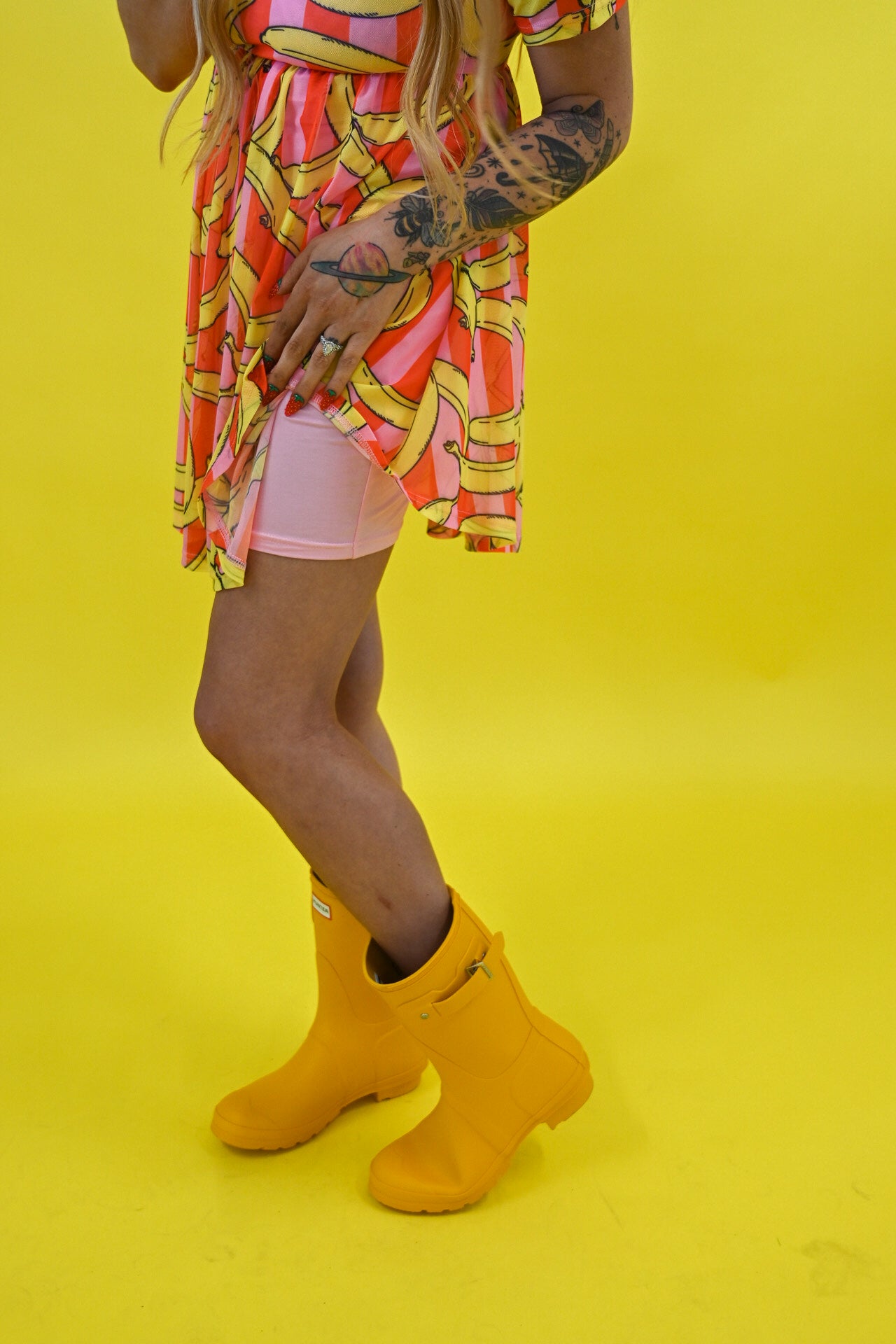 A person wearing a bright yellow rain boot with a small white brand tab, paired with a pink and yellow patterned dress and light pink shorts, standing against a solid bright yellow background, close up side view