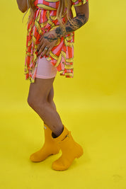 A person wearing a bright yellow rain boot with a small white brand tab, paired with a pink and yellow patterned dress and light pink shorts, standing against a solid bright yellow background, close up side view