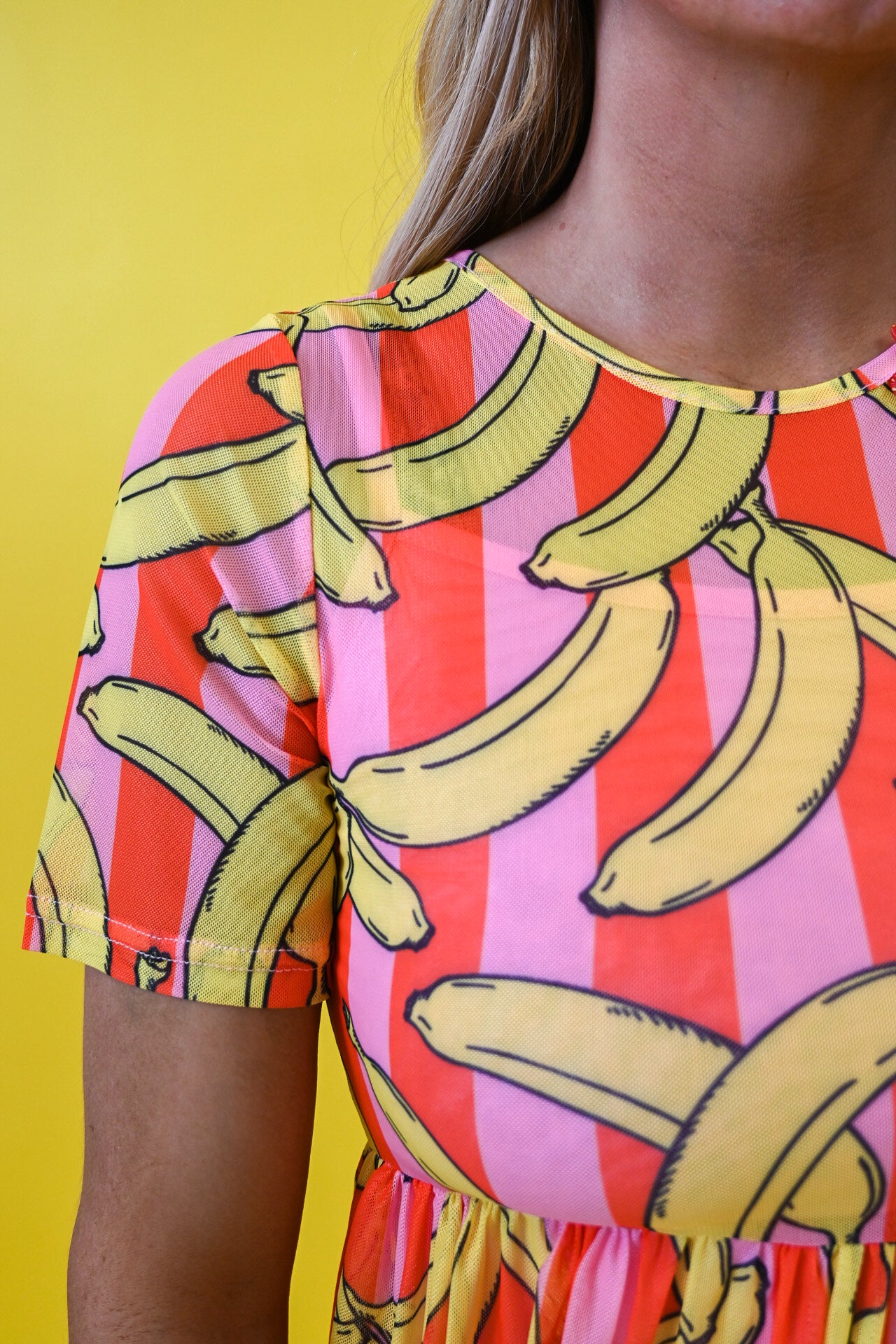 A close-up of a person wearing a short-sleeve top with a bold banana print over vertical pink, red, and orange stripes, set against a solid yellow background 