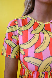 A close-up of a person wearing a short-sleeve top with a bold banana print over vertical pink, red, and orange stripes, set against a solid yellow background 
