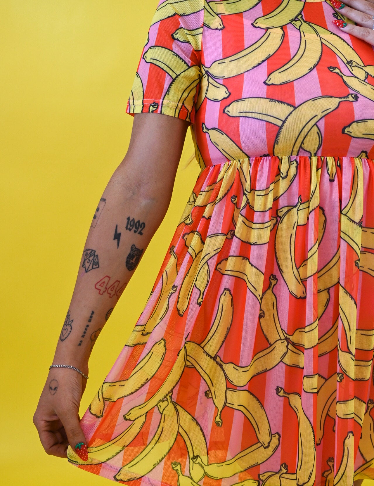 A woman wearing a short-sleeve dress with a vibrant yellow banana print over pink and orange striped vertical bands, posed against a solid yellow background