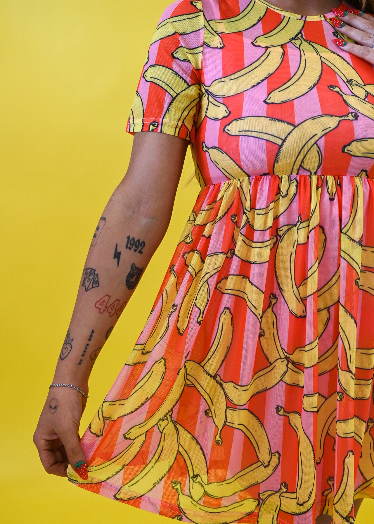 A woman wearing a short-sleeve dress with a vibrant yellow banana print over pink and orange striped vertical bands, posed against a solid yellow background