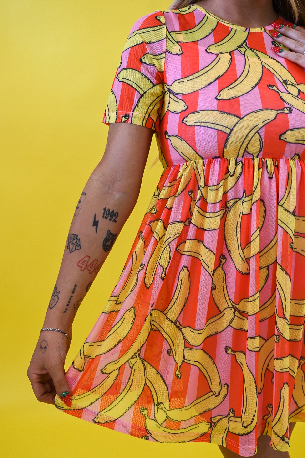 A woman wearing a short-sleeve dress with a vibrant yellow banana print over pink and orange striped vertical bands, posed against a solid yellow background