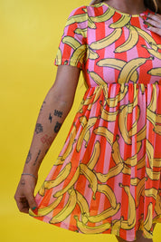 A woman wearing a short-sleeve dress with a vibrant yellow banana print over pink and orange striped vertical bands, posed against a solid yellow background