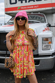 A woman stands in front of a white truck wearing a bright pink beanie, black sunglasses, a pink and yellow banana-print mini dress, and a leopard-print crossbody bag, close up view