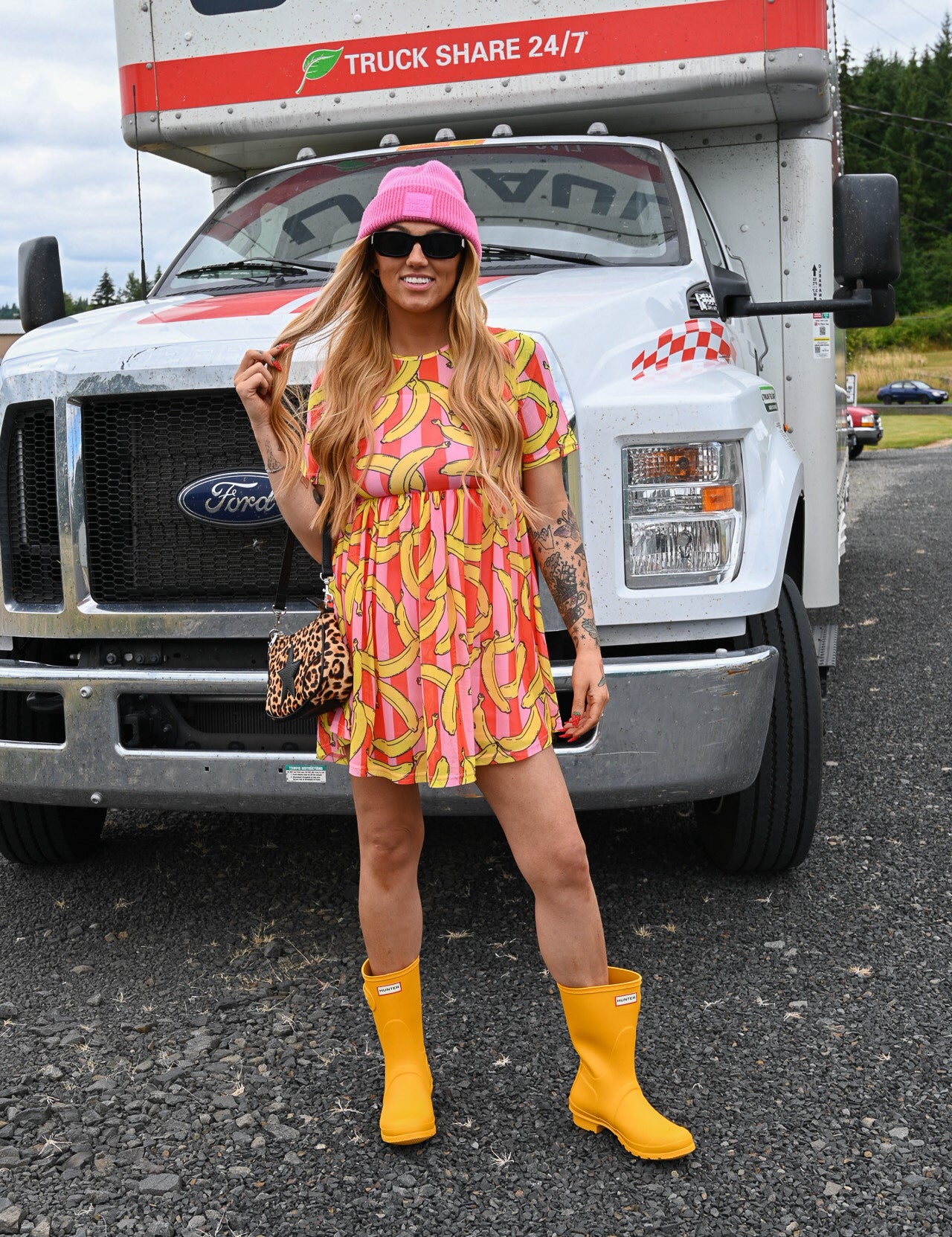 A woman stands in front of a large white U-Haul truck wearing a pink and yellow banana-print mini dress, a pink bucket hat, yellow rain boots, and a leopard-print handbag