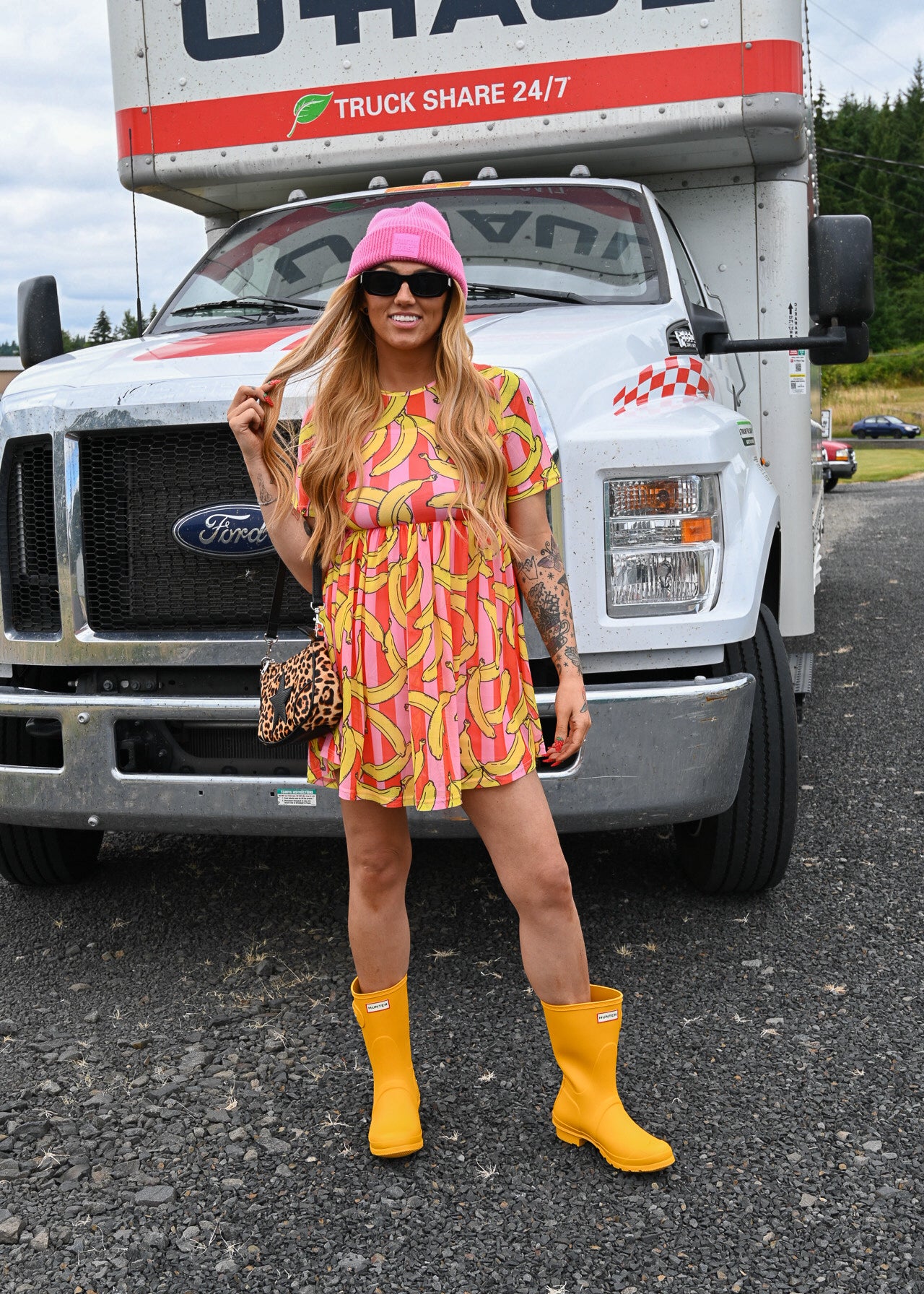 A woman stands in front of a large white U-Haul truck wearing a pink and yellow banana-print mini dress, a pink bucket hat, yellow rain boots, and a leopard-print handbag