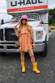 A woman stands in front of a large white U-Haul truck wearing a pink and yellow banana-print mini dress, a pink bucket hat, yellow rain boots, and a leopard-print handbag