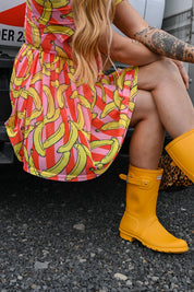 A person seated outdoors wearing a bright dress with an allover banana print in yellow, pink, and orange, paired with bright yellow rain boots. The photo is cropped to show the lower torso, legs, and boots, with gravel on the ground and part of a vehicle in the background
