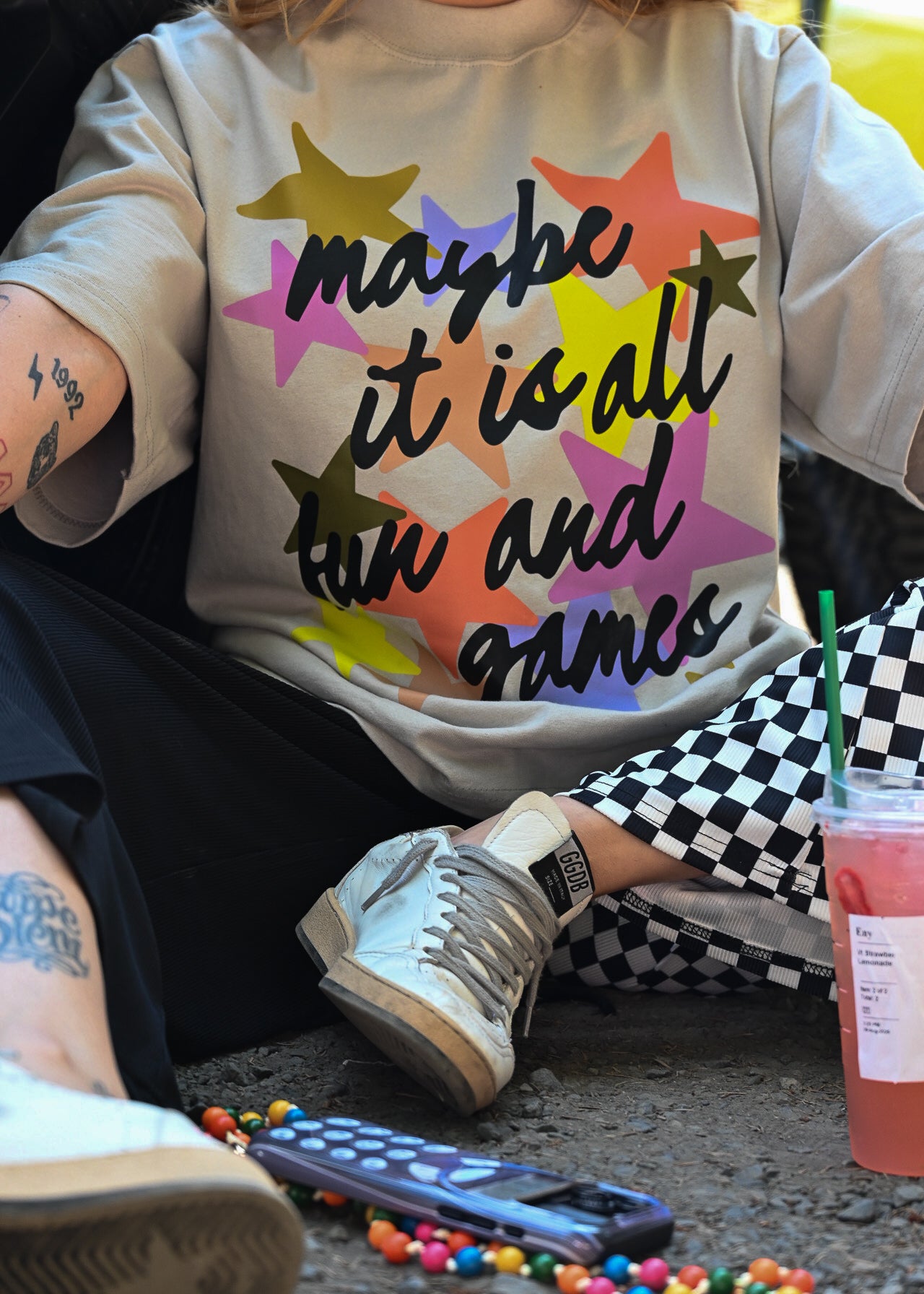 A person wearing a light beige graphic t-shirt with the text 'maybe it is all fun and games' in black script over a colorful pattern of overlapping stars. The person is sitting outdoors on a concrete surface, wearing black and white checkered pants, white sneakers, and holding a pink drink