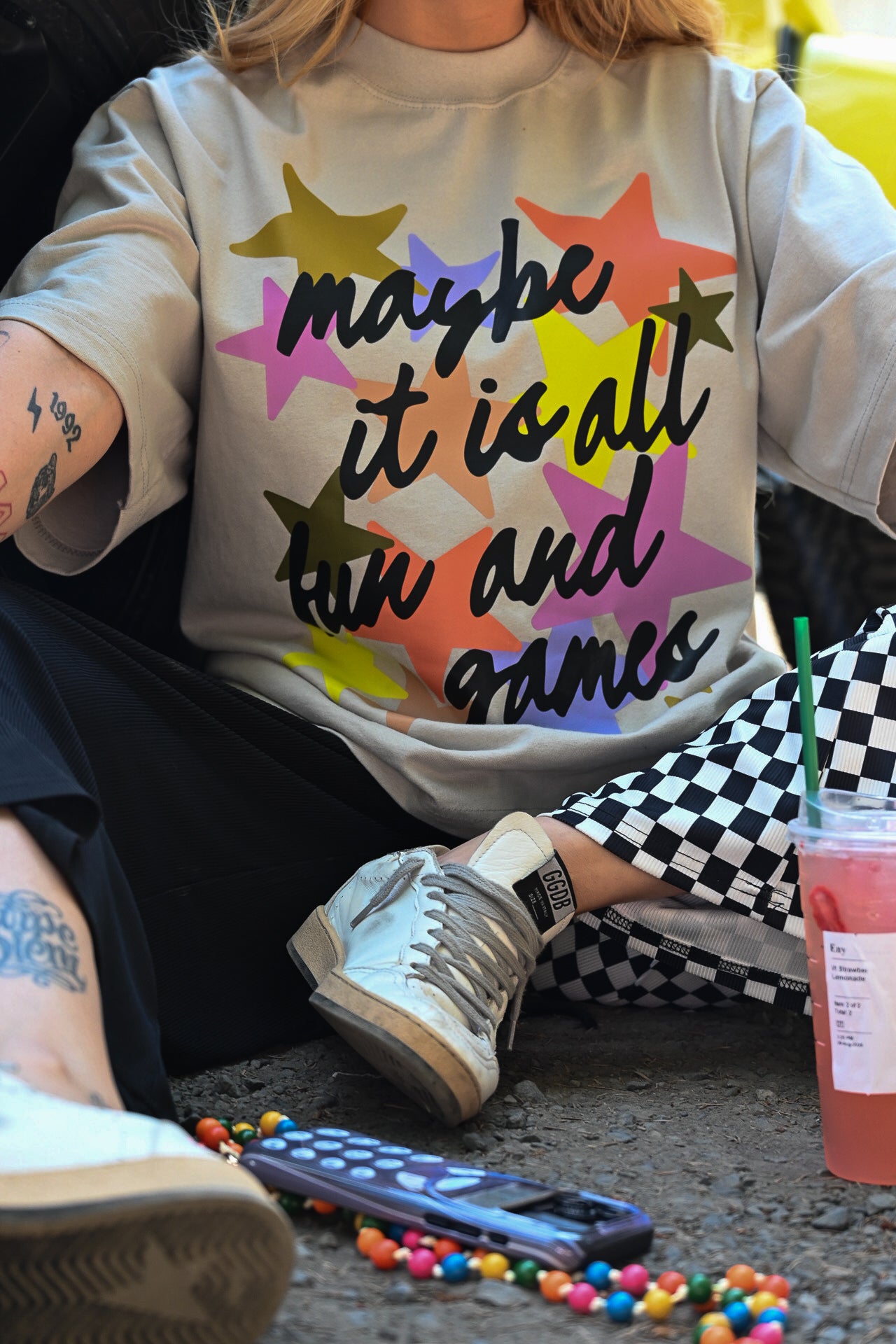 A person wearing a light beige graphic t-shirt with the text 'maybe it is all fun and games' in black script over a colorful pattern of overlapping stars. The person is sitting outdoors on a concrete surface, wearing black and white checkered pants, white sneakers, and holding a pink drink