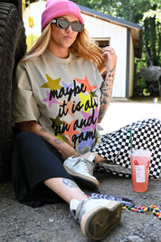 A woman sitting outdoors on gravel next to a tire, wearing a beige graphic t-shirt with the text 'maybe it is all fun and game!' in colorful star shapes, a pink beanie, black-and-white checkered pants, and white sneakers. A pink drink is on the ground nearby