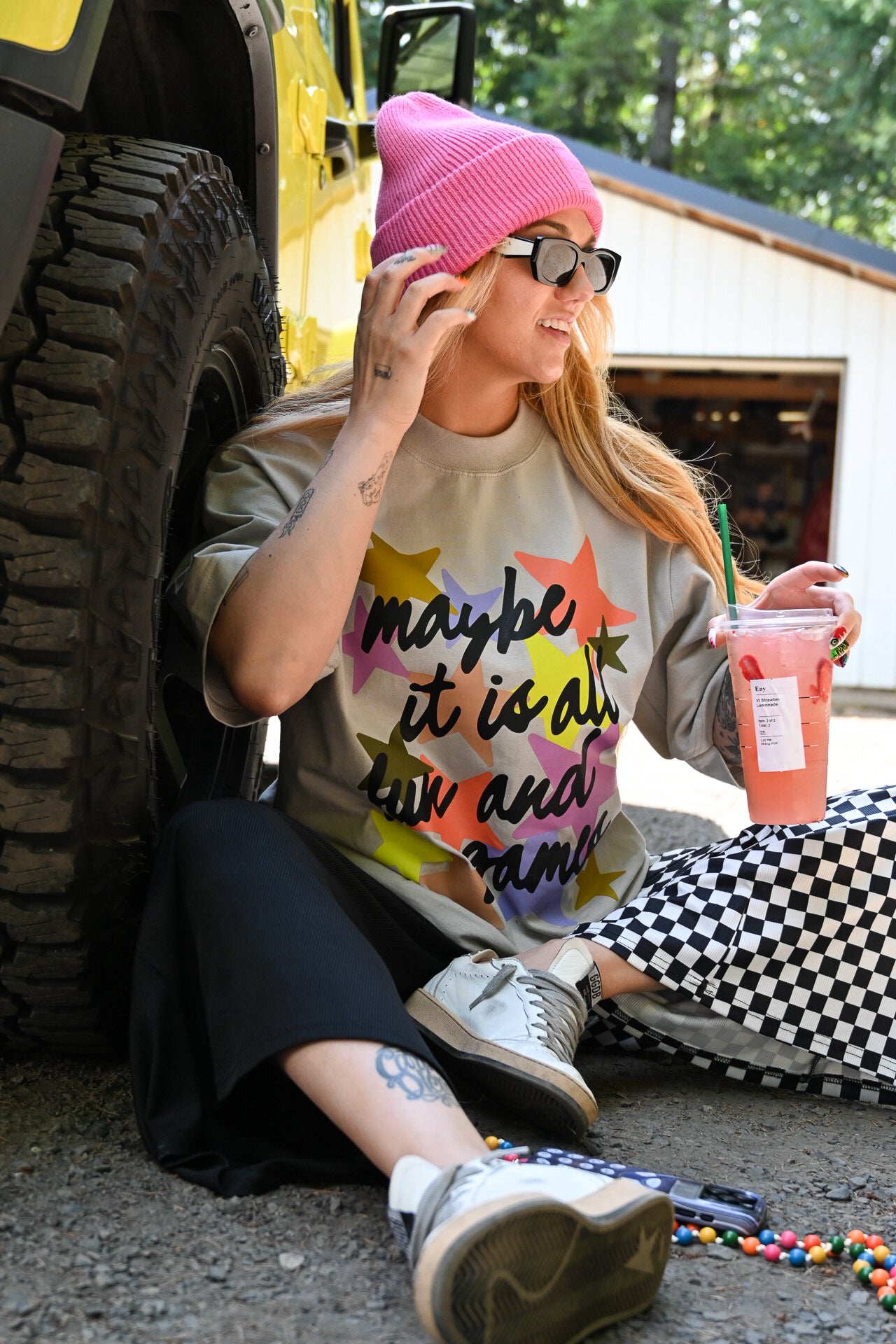 A woman sitting on the ground next to a yellow vehicle, wearing a pink beanie, black sunglasses, a graphic t-shirt with colorful star shapes and the text 'maybe it is all fun and games', black pants, and white sneakers. She is holding a pink drink