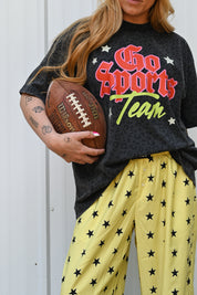 A person wearing a black graphic T-shirt that reads 'Go Sports Team' in red, orange, and green, paired with yellow pants covered in black stars, holding a brown football against a light gray wall